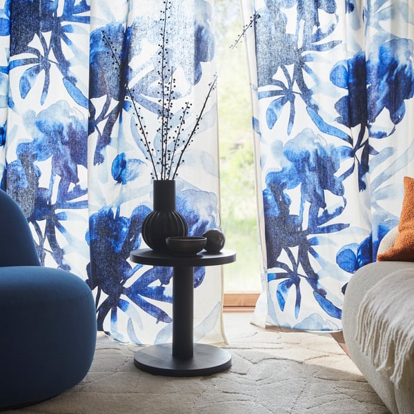 Sala con cortinas SKOGSNÄSSLA blancas y azules translúcidas con diseño floral, junto a un sillón azul y una mesa.