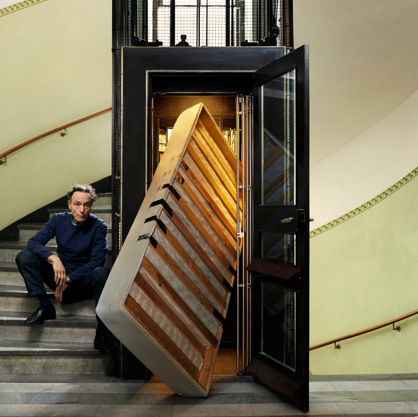 A person sits on a staircase next to a bed base stuck in the elevator—an illustration of a common problem that a removal service can easily solve.