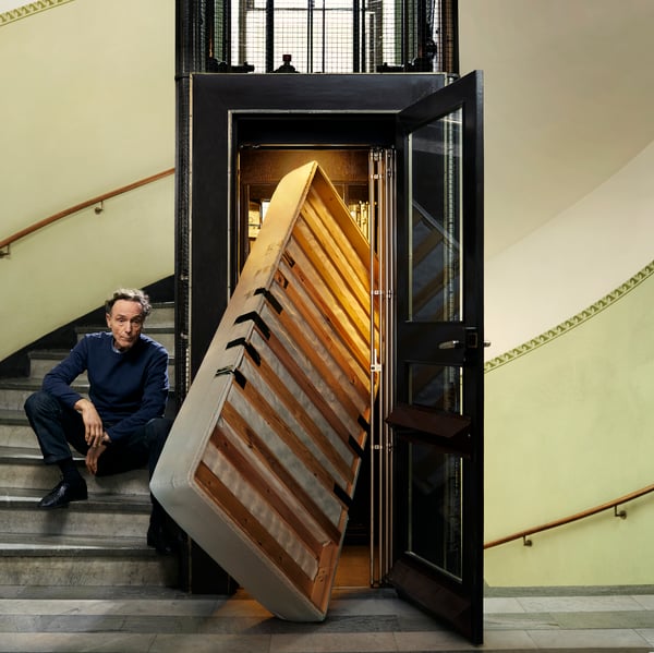 A person is sitting on a curved staircase next to an elevator where an old mattress protrudes through the open door.