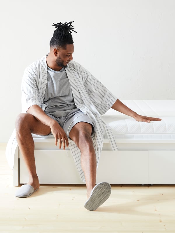 A person sits at the end of a white MALM bed with 2 storage boxes pressing down on an ÅKREHAMN foam mattress with one hand.
