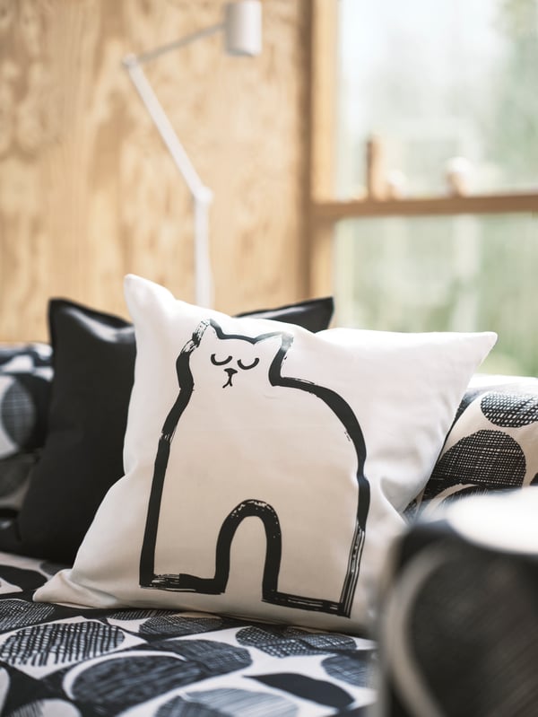 A cushion with the TALLSPINNARE cushion cover is on a soft bed. The cover is decorated with an outline of a cat.