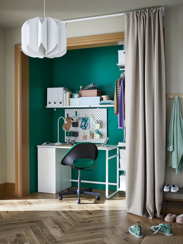 Un escritorio blanco ubicado en un rincón con muros verdes, junto al vestíbulo de una casa o departamento con piso de parquet. Frente al escritorio hay una silla de altura ajustable con asiento verde, y sobre el escritorio una repisa con artículos de trabajo. 