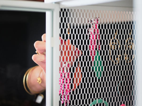 The hand of a person wearing gold bracelets and rings opening the white mesh metal door of a BAGGEBO cabinet.