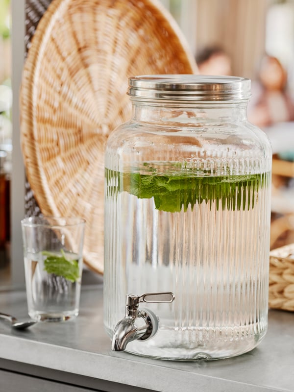 Großes Glasgefäß mit Gurkenwasser und Auslaufhahn auf einer Küchenarbeitsplatte, daneben ein Glas mit Gurkenscheiben – erfrischendes Getränk in stilvoller Umgebung.