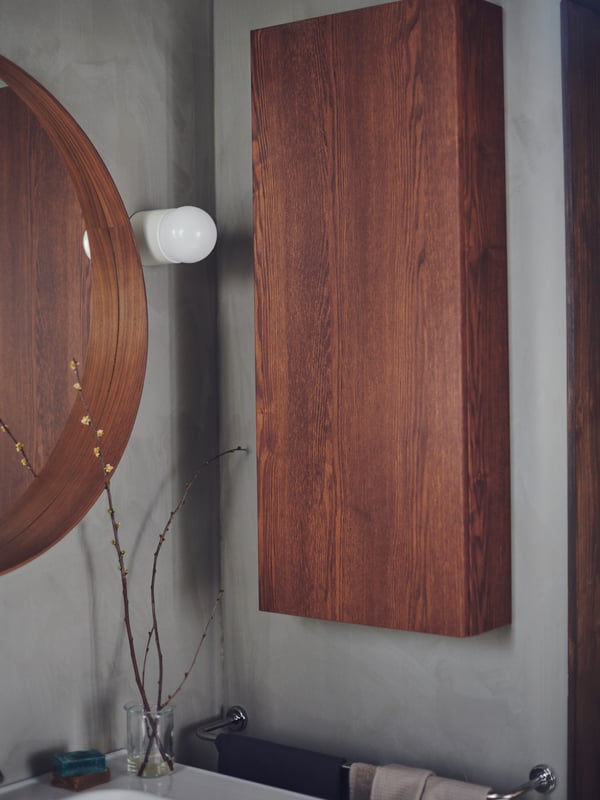A wood-finish GODMORGON cabinet on a bathroom wall beside a STOCKHOLM mirror and with a wash-basin and towel rail below it.