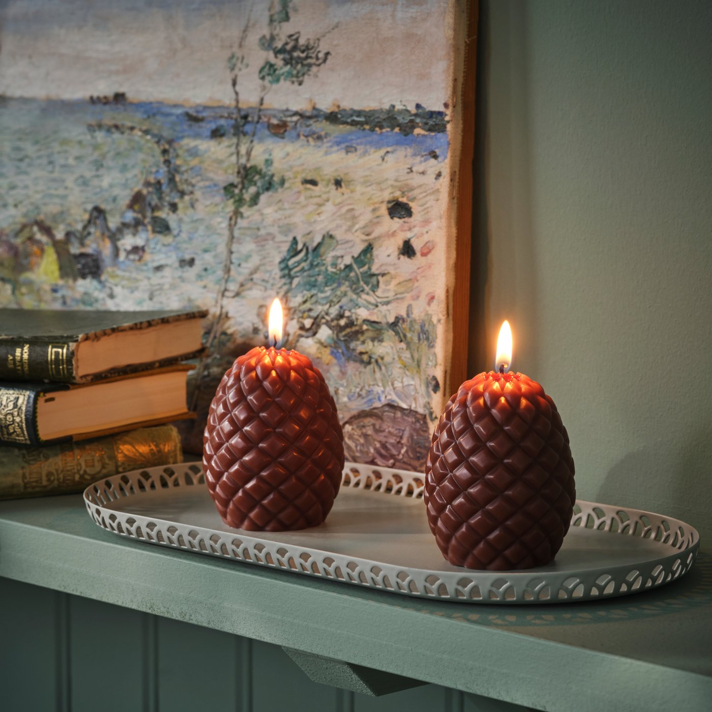 A couple of SNÖSISKA pillar candles shaped like pine cones stand on a pale grey-green SNÖSISKA candle dish on a green shelf.