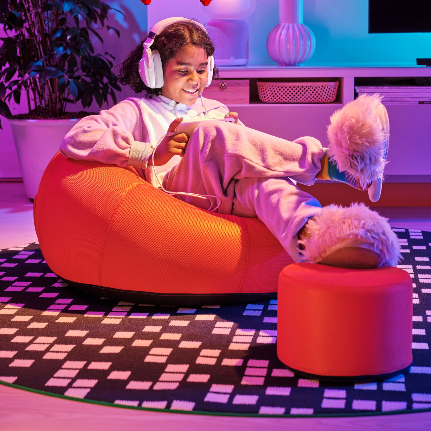 Person i hörlurar sitter i orange sittsäck med fotpall som står på en BRÄNNBOLL svart och vit matta. Personen har fluffiga tofflor och det är färgglad belysning i rummet.