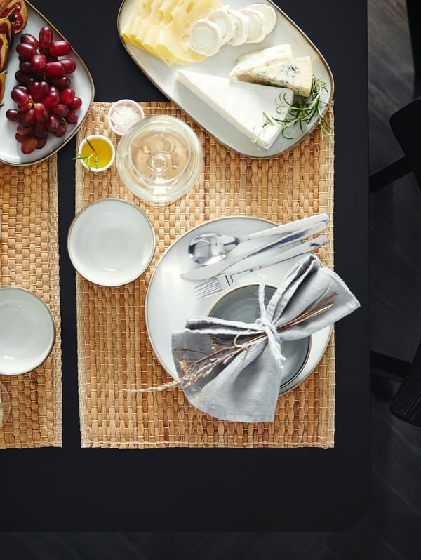 Ein Tisch mit Tischsets aus Wasserhyazinthe, grauem Geschirr uns zwei Servierplatten mit Käse und Trauben.