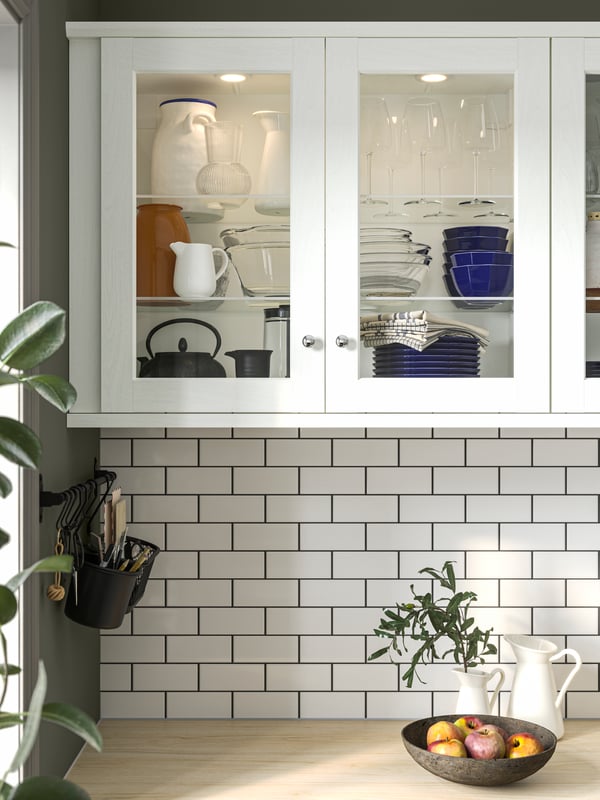 Glass cabinets with white ENKÖPING frames have kitchenware stacked on shelves, illuminated by MITTLED spots on the inside.