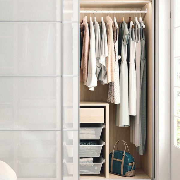 An open PAX wardrobe with white sliding doors has a white-stained oak-patterned interior with clothes, baskets, and drawers.