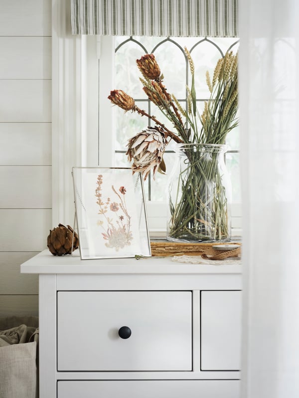 The top left side of a white HEMNES chest with 8 drawers that has a vase of dried flowers and dried flower picture on top.