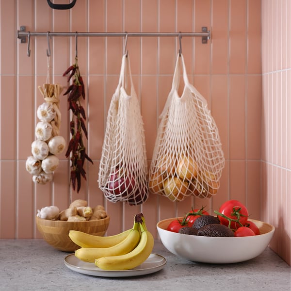 Fruits et légumes disposés dans des bols sur un comptoir gris, et suspendus dans des sacs en filet ainsi qu’aux crochets d’une barre KUNGSFORS en acier inoxydable.