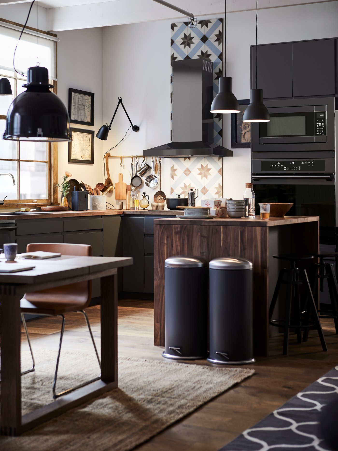 A black kitchen with a cosy lighting plant. On the floor, there's a natural LOHALS rug, flatwoven.