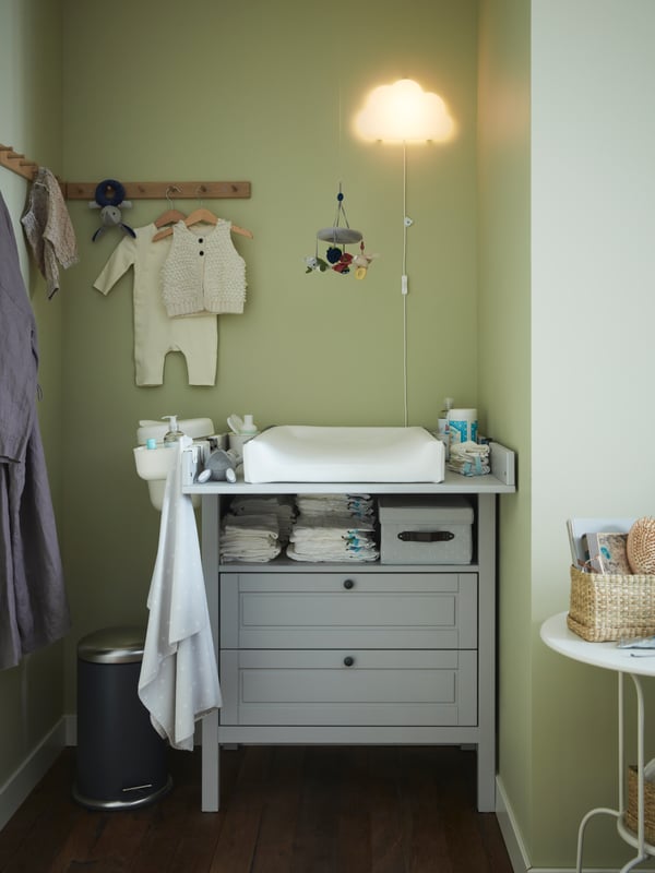 Un mudador SUNDVIK gris claro cubierto de implementos para mudar en una pieza con muros verde claro. En el muro hay una lámpara de noche con forma de nube UPPLYST y un colgador de madera HÖVOLM para su ropa.