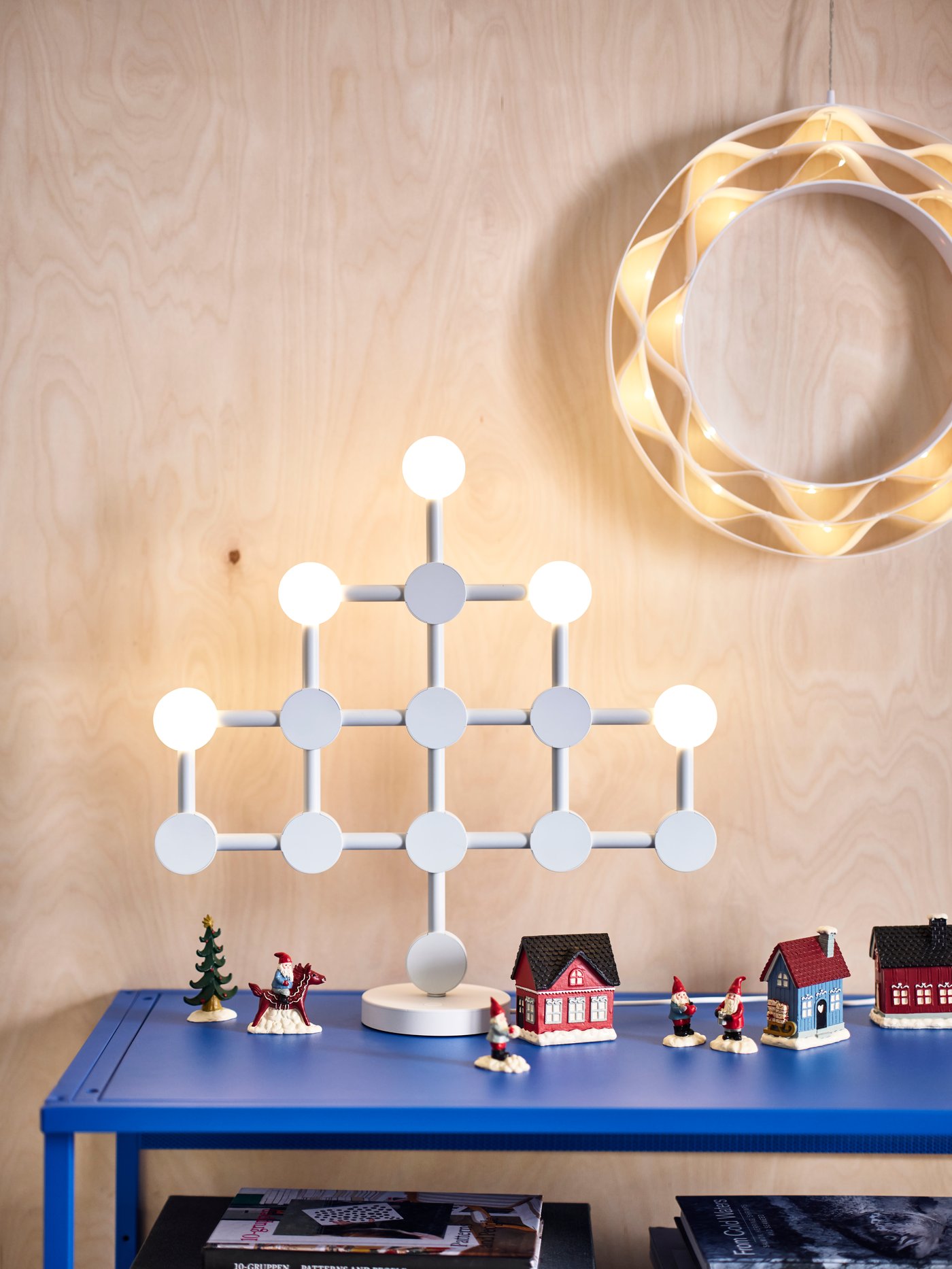 A modern, white STRÅLA LED table lamp sits on a table among a VINTERFINT winter village decoration with gnomes and houses.