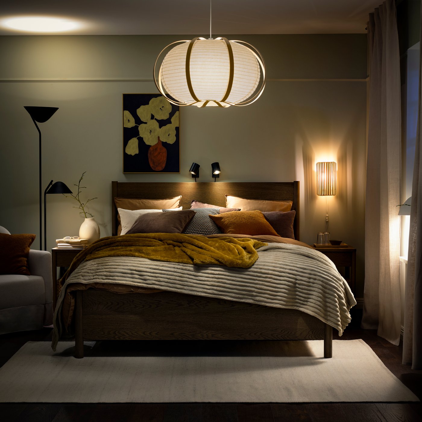 A bedroom with cosy lighting from TAGGTOFS lamp shade and a dark brown TONSTAD bed covered in throws and pillows.