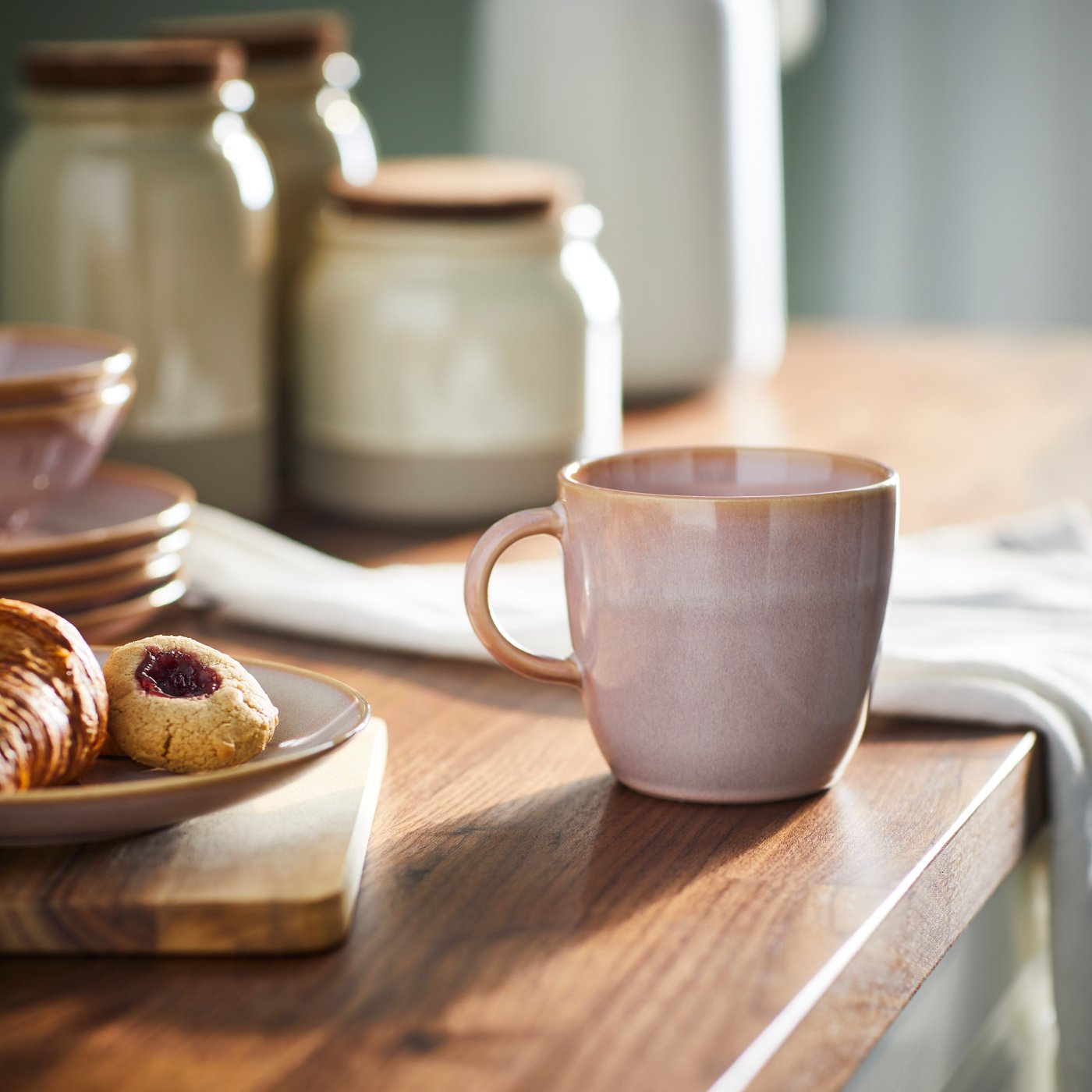 A light pink FÄRGKLAR mug stands on a surface in a kitchen near a FÄRGKLAR plate with some food on it.