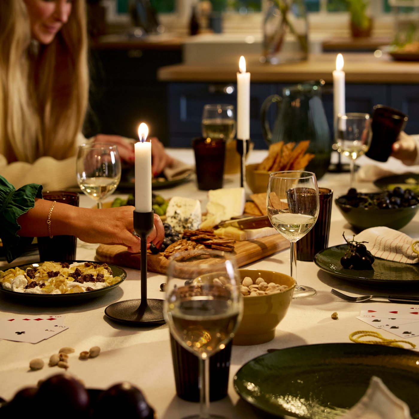 People are playing cards at a festive dinner table with candle sticks, wine glasses and plates with cheese and crackers.