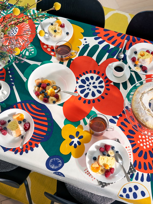 Table with bright colourful tablecloth with flower print, white plates and bowls with breakfast foods, and black chairs.