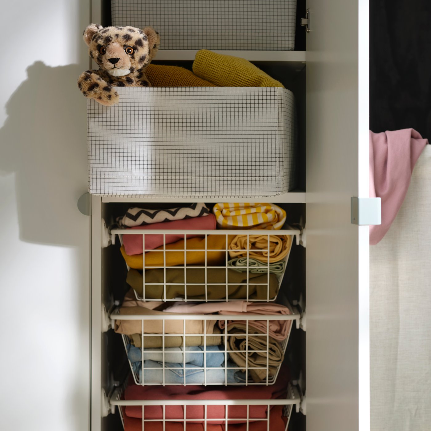 Una caja de almacenaje STUK en un clóset, llena de ropa doblada y un peluche en la parte superior, junto a varias prendas colgadas. 
