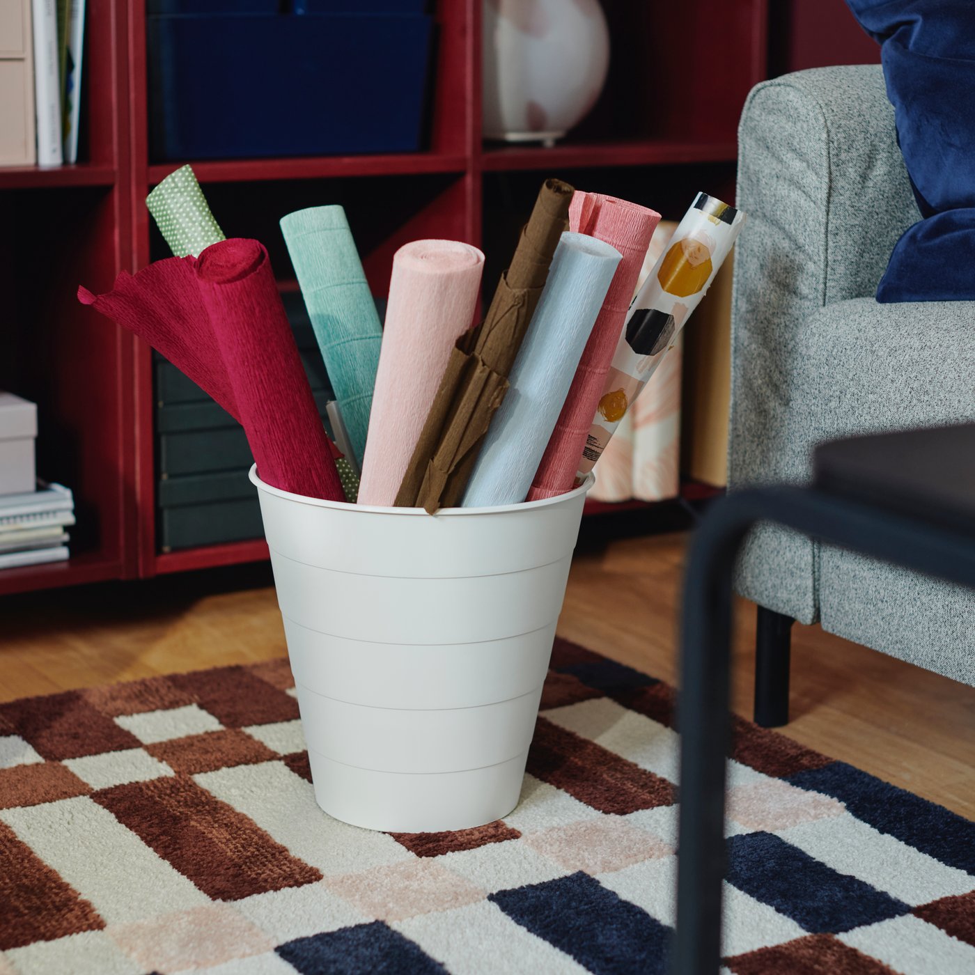 A white FNISS waste bin with rolls of coloured paper in it stands on a MARKBLÅMME rug in front of red shelving units.