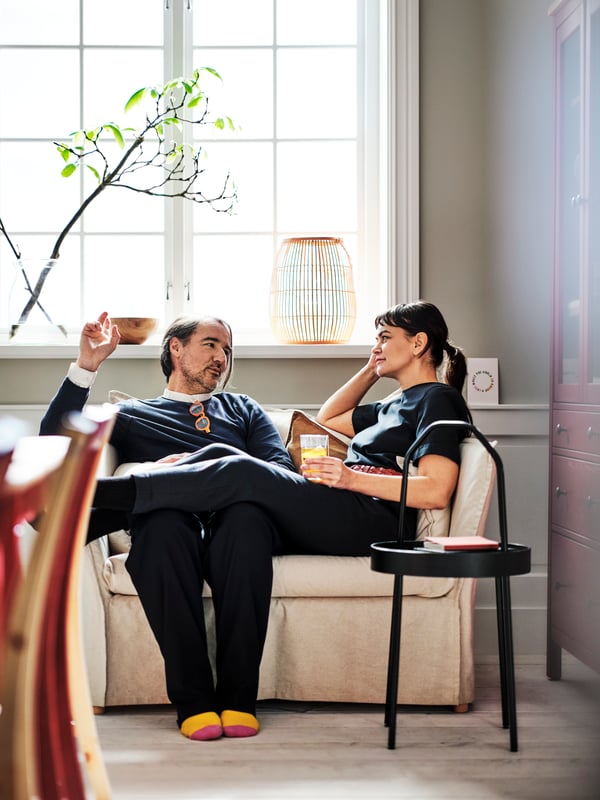 Two people chatting while seated on a natural BACKSÄLEN 1,5-seat armchair near a black BURVIK side table.