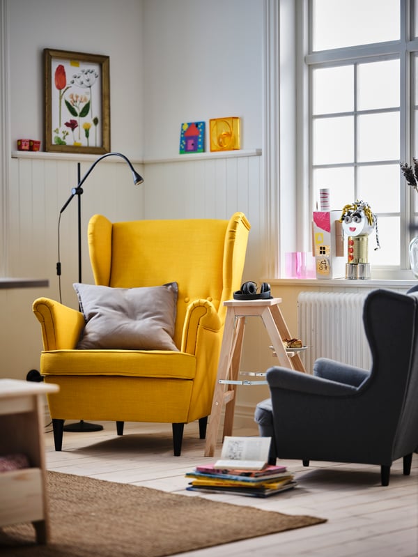 A living-room reading corner with a yellow STRANDMON wing chair and a grey STRANDMON children’s armchair.
