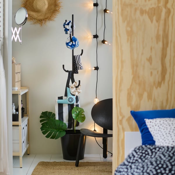A black ÄLGANÄS hat and coat stand holds hair accessories with a SVARTRÅ LED light chain and a FRÖSET easy chair beside it.