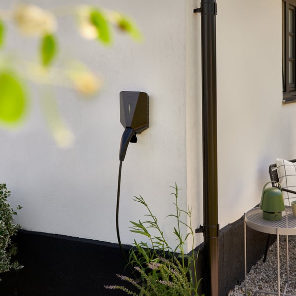 Une station de recharge pour véhicules électriques installée à l’extérieur d’une maison. En haut à gauche de l’image, une branche avec des feuilles vertes apparaît floue.