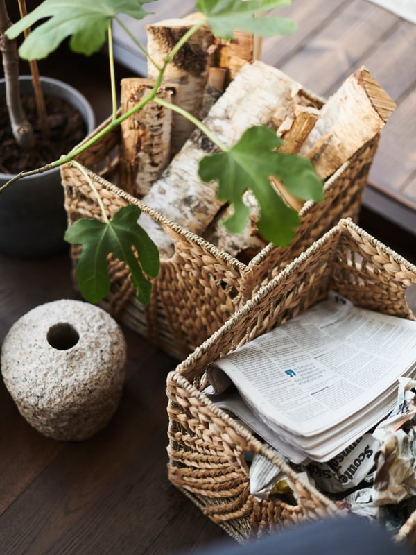 Two natural-colored LUSTIGKURRE baskets on the floor by a plant. One is filled with newspaper and the other holds logs.