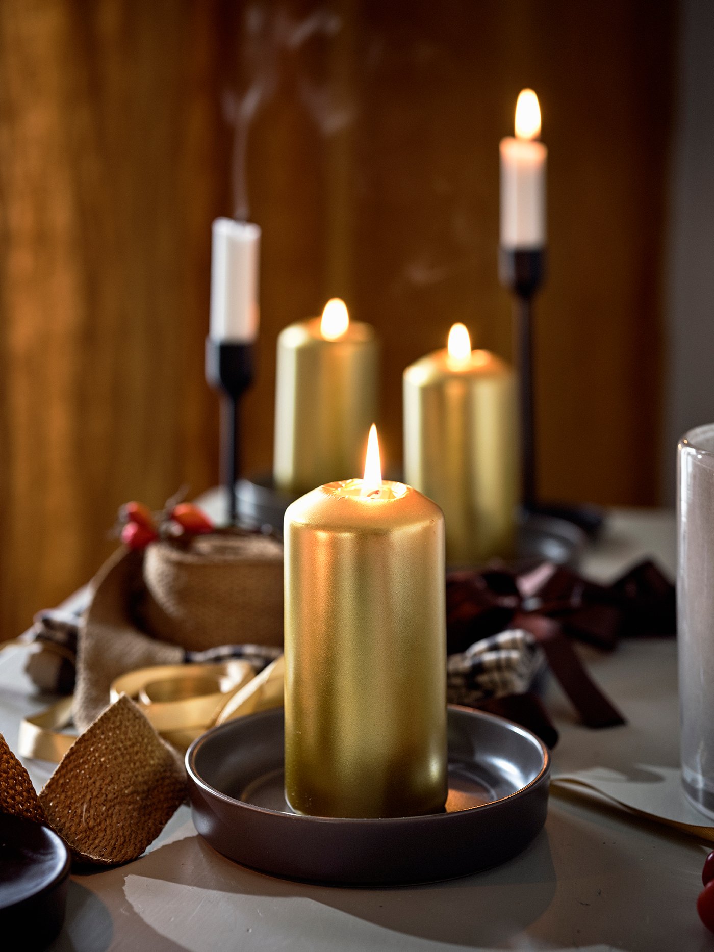 Sobre una mesa hay velas FENOMEN doradas; al fondo se ven velas altas en candelabros negros.