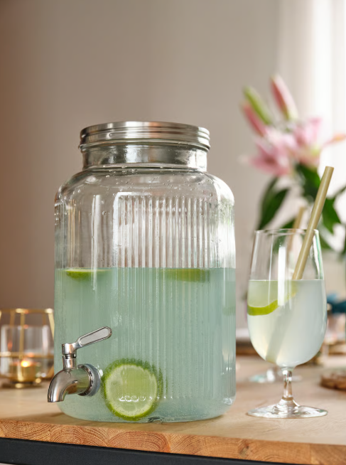 Grosser Getränkespender mit grünem Zitronengetränk und Limettenscheibe auf Tisch, dahinter Glas und Blumenarrangement.