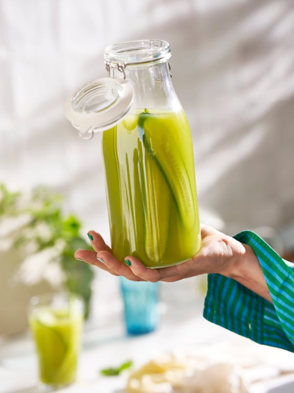 A person is holding a KORKEN bottle shaped jar with the lid open. The bottle is filled with a green-coloured water infusion.