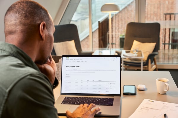 ラップトップでIKEA Business Networkの取引履歴ページを開き、テーブル上の書類やスマートフォン、コーヒーカップとともに確認する人。