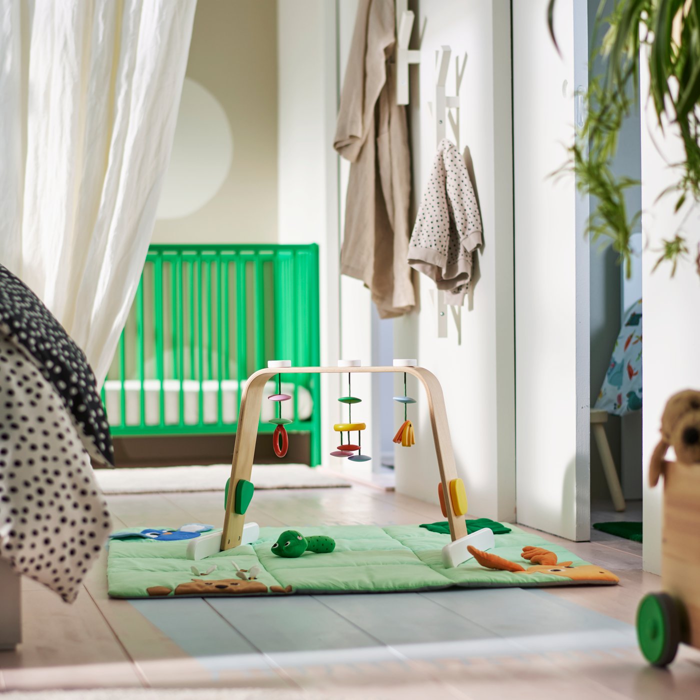 A multicoloured LEKA baby gym with hanging toys on a green playmat on the floor in a bedroom with a green GULLIVER cot.