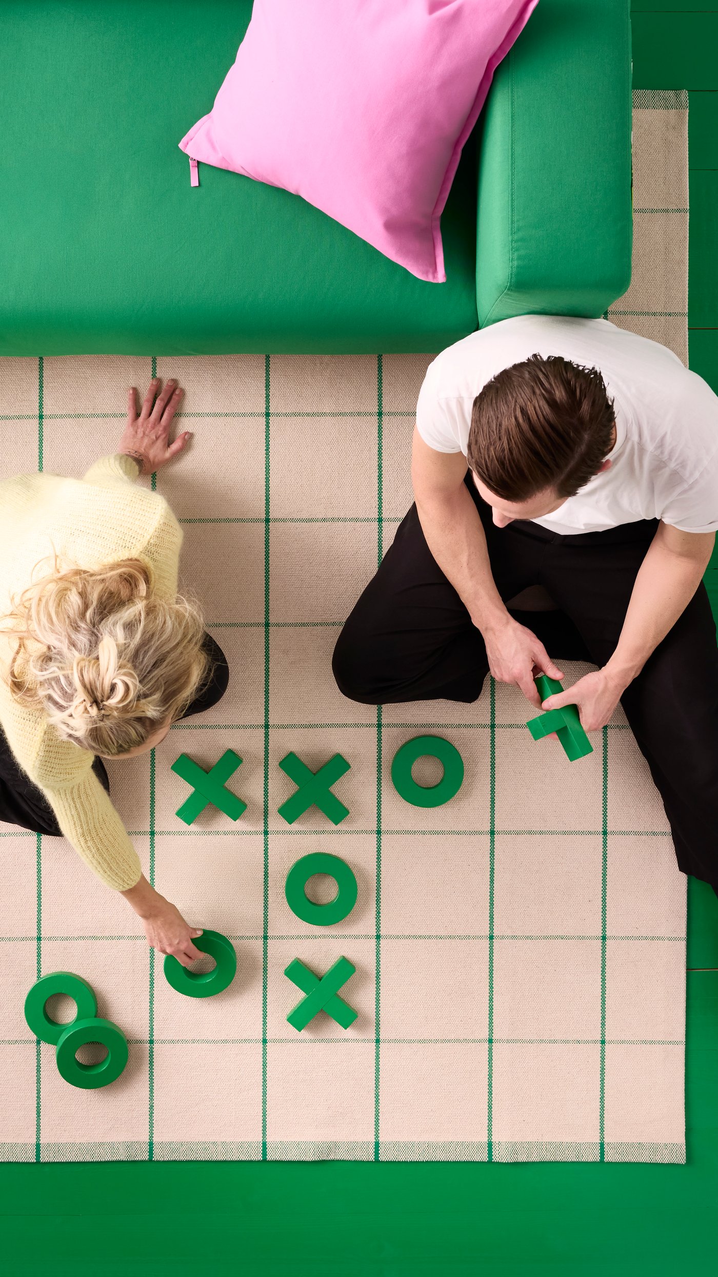 En rutig matta med ett stort spelplanmönster där två personer spelar fyra i rad på mattans rutor. En grön soffa syns delvis i bildens övre kant.