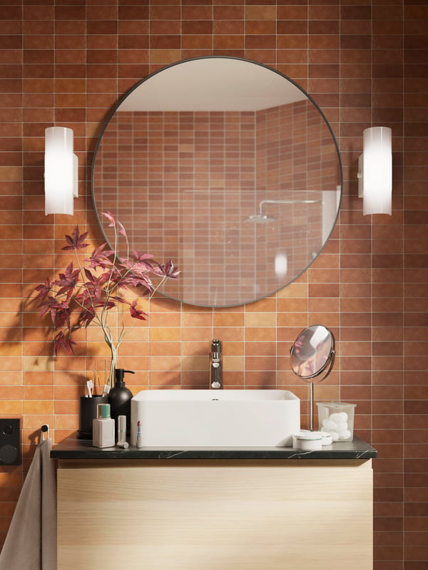 A round LINDBYN mirror is mounted above a white sink in a bathroom with brown tiles. There are white lamps on either side.