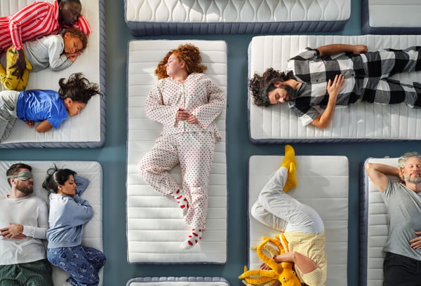 Et foto set ovenfra af ni personer i forskellige aldre, der sover på forskellige madrasser, som ligger på et gråt gulv i et atelier.