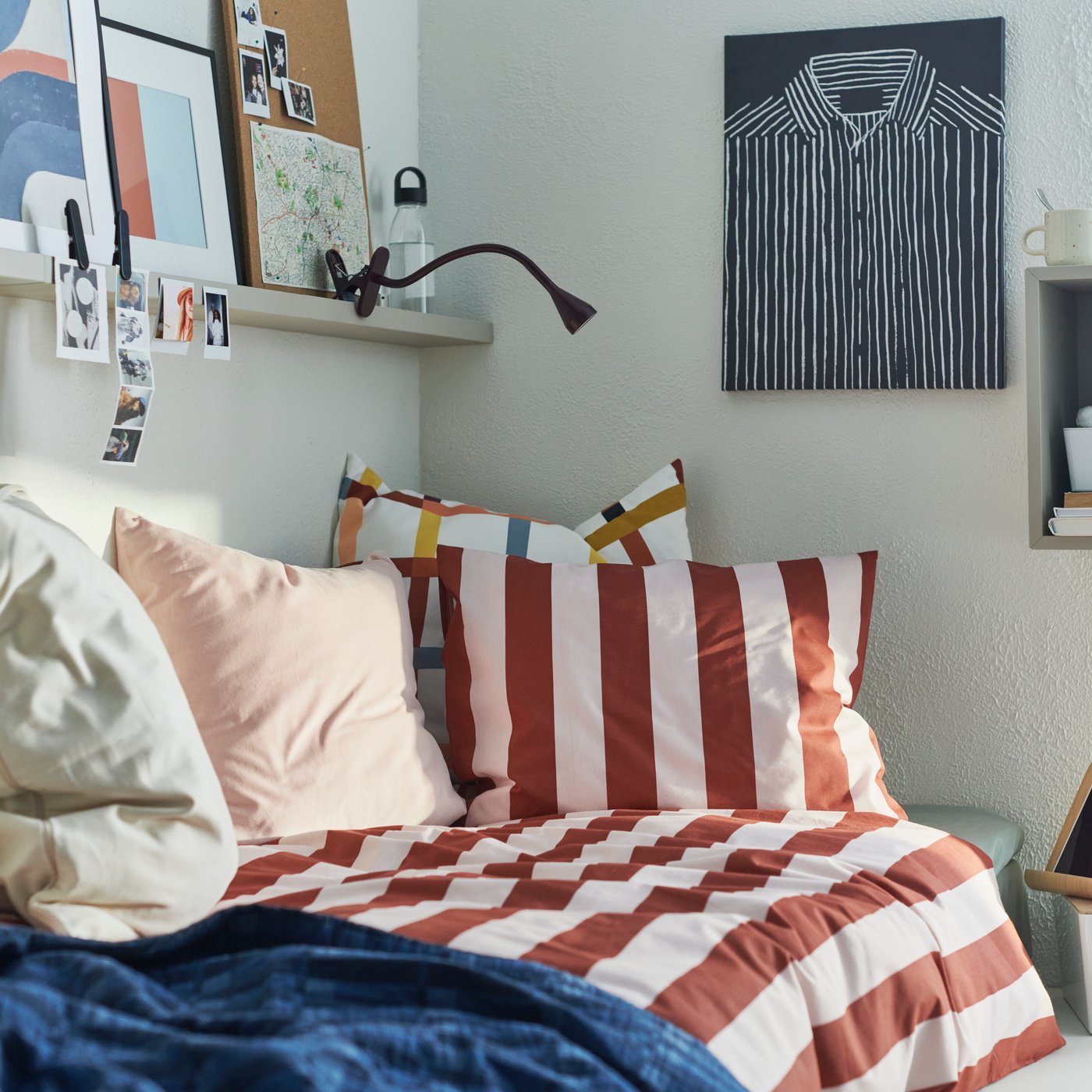 A dorm room with a light pink and dark red-striped SLÅNHÖSTMAL duvet cover and pillowcase on a bed with cushions.