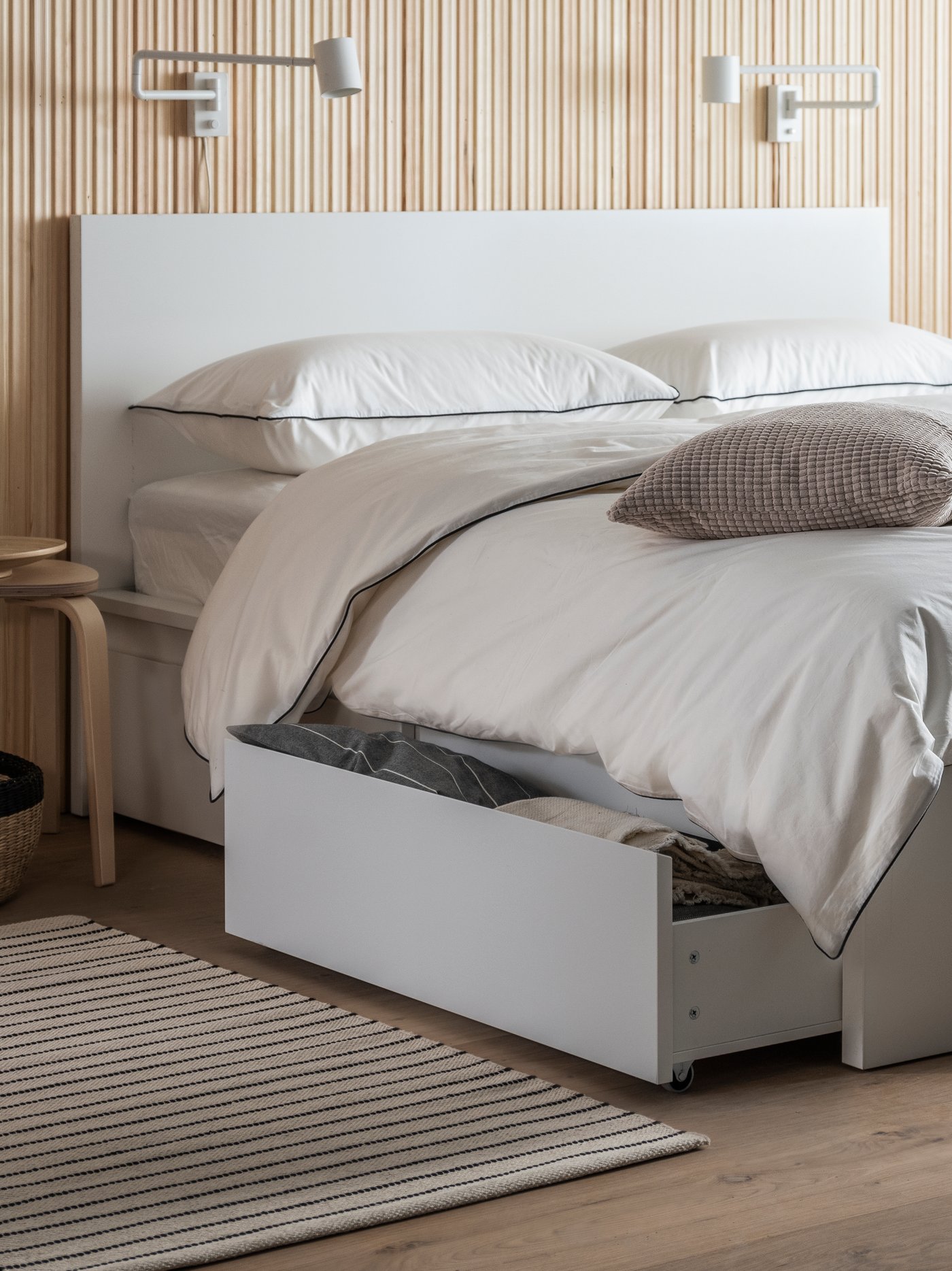 A bedroom setting with a bed and opened storage drawer. A white and black rug has been placed to the side of the bed.
