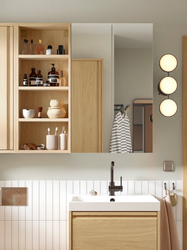 Un baño con gabinetes en color madera del sistema ÄNGSJÖN, un espejo en la pared y al costado un mueble de baño mural.