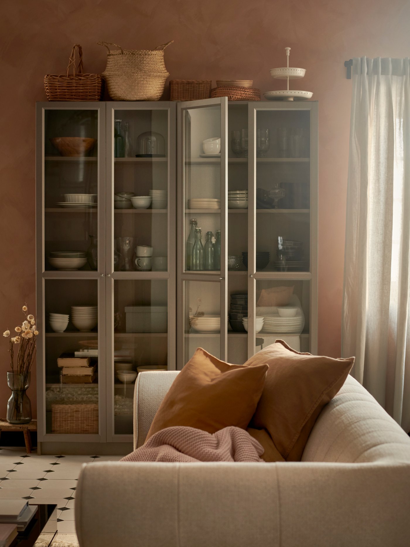 Et BILLY skab med glaslåger står bagerst i en stue foran står en hvid stof sofa med terracotta farvede puder.