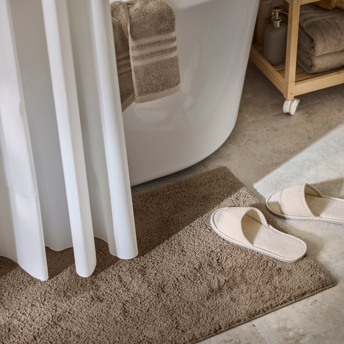A bathroom floor with a BACKMÅRA bath mat next to a a white bath tub with a white shower curtain and a pair of slippers.