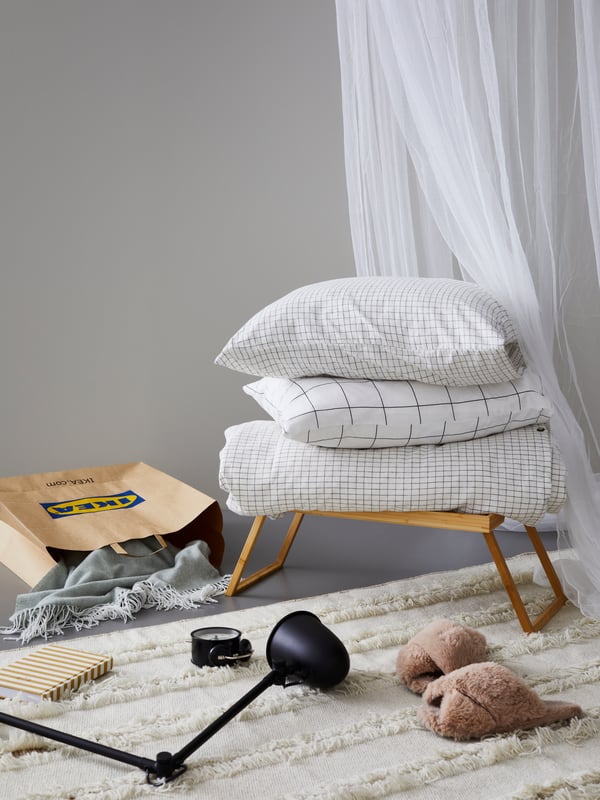ÅKERFIBBLA bed linen is on a RESGODS tray table. An IKEA bag, HOLMVI throw, SKURUP work/wall lamp and DEKAD clock are nearby.