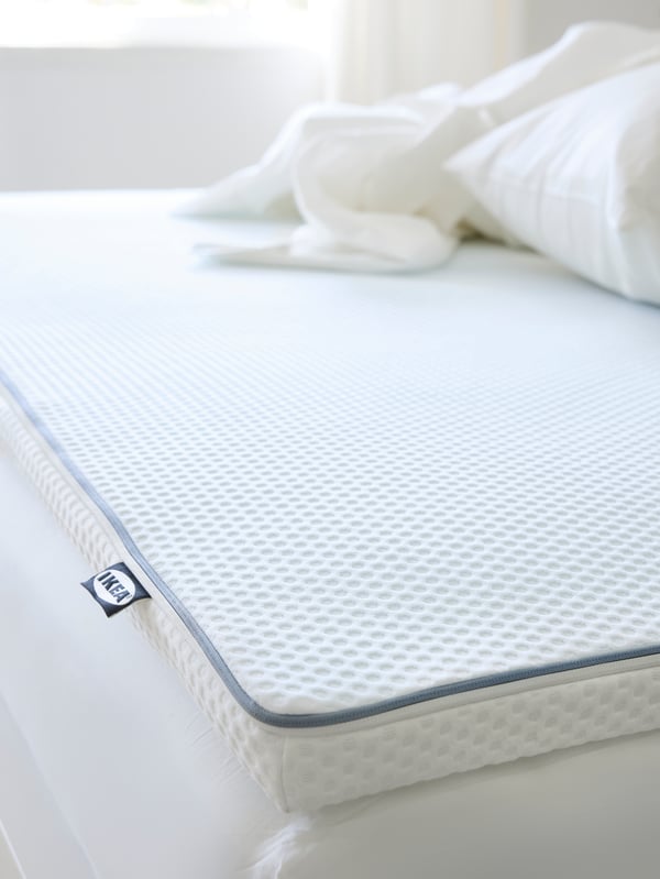 Close-up of NÄSTABERG mattress pad in a cooling and moisture wicking material. White pillow and quilt in a pile on top.