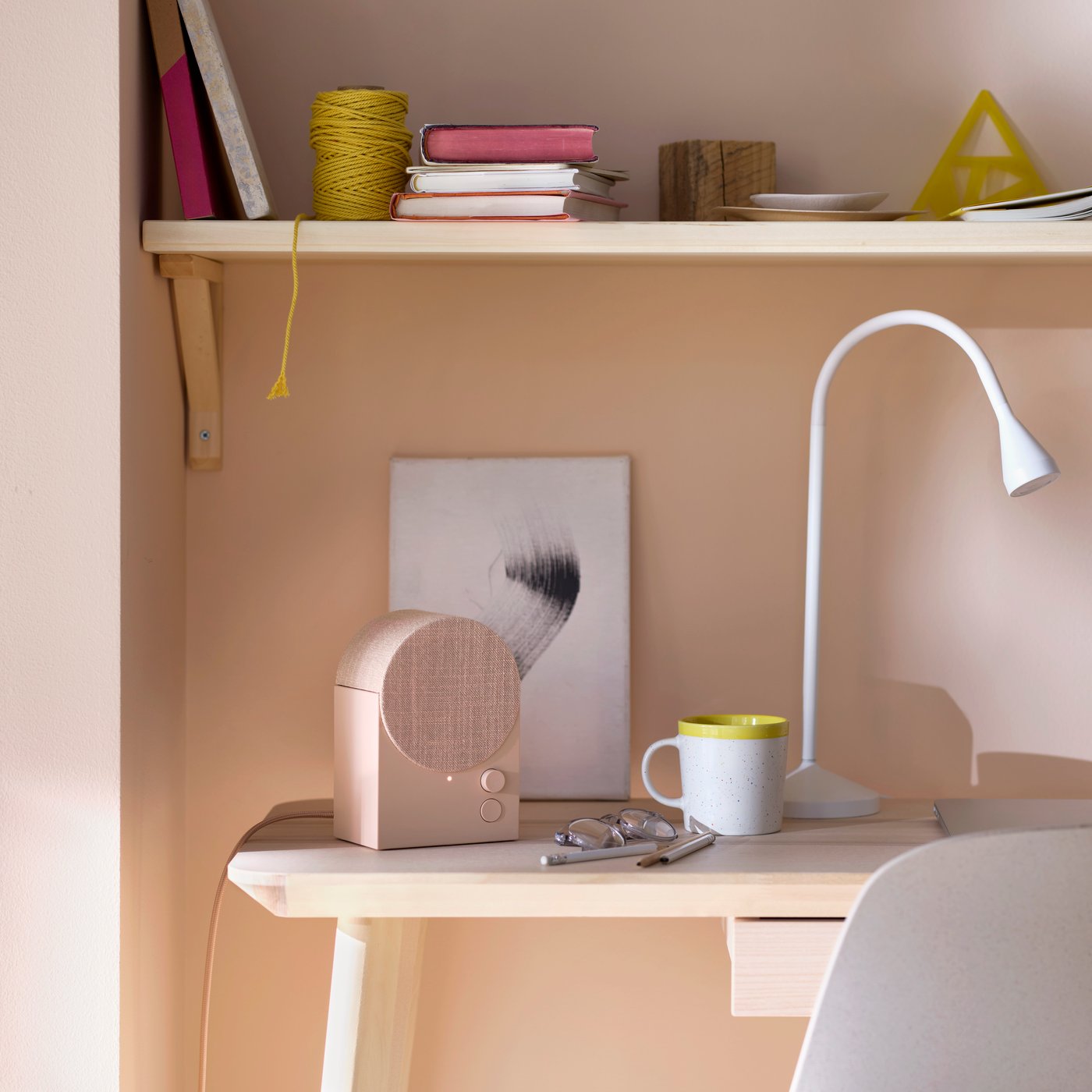 A NATTBAD Bluetooth speaker, a white NÄVLINGE work lamp and a mug stand on a desk underneath a wall shelf.
