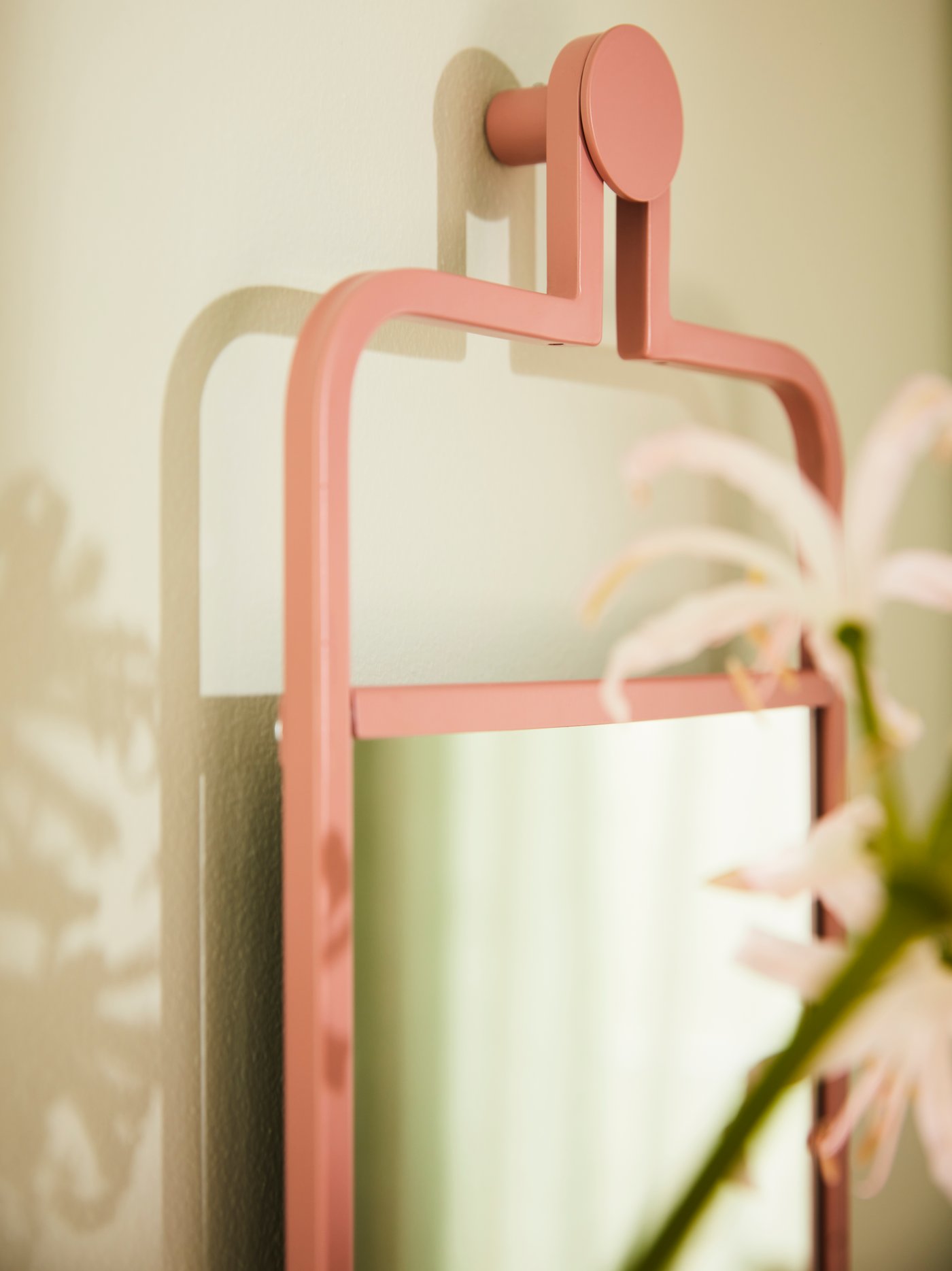 Un miroir rose est accroché à un mur couleur crème et forme une ombre derrière. Des lys flous apparaissent au premier plan.
