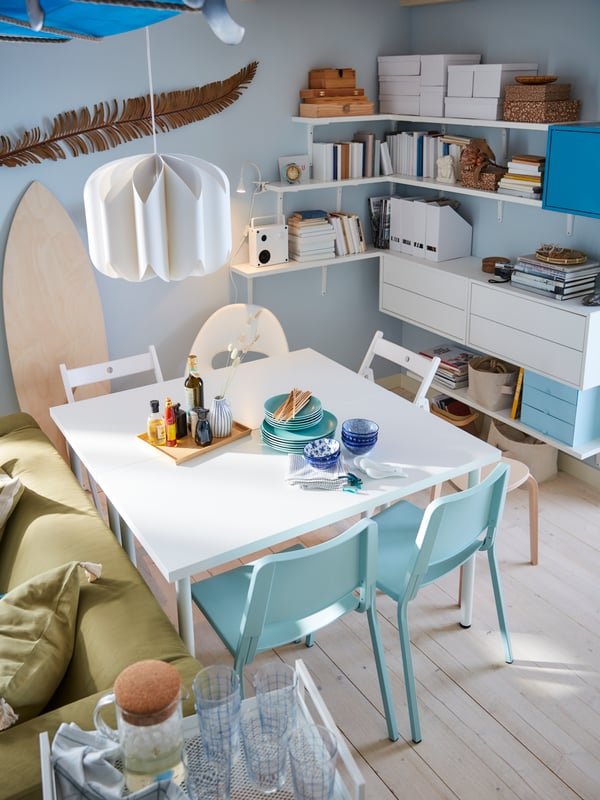A white dining table, light turquoise and white chairs, white pendant lamp, blue walls, white shelves and brackets.