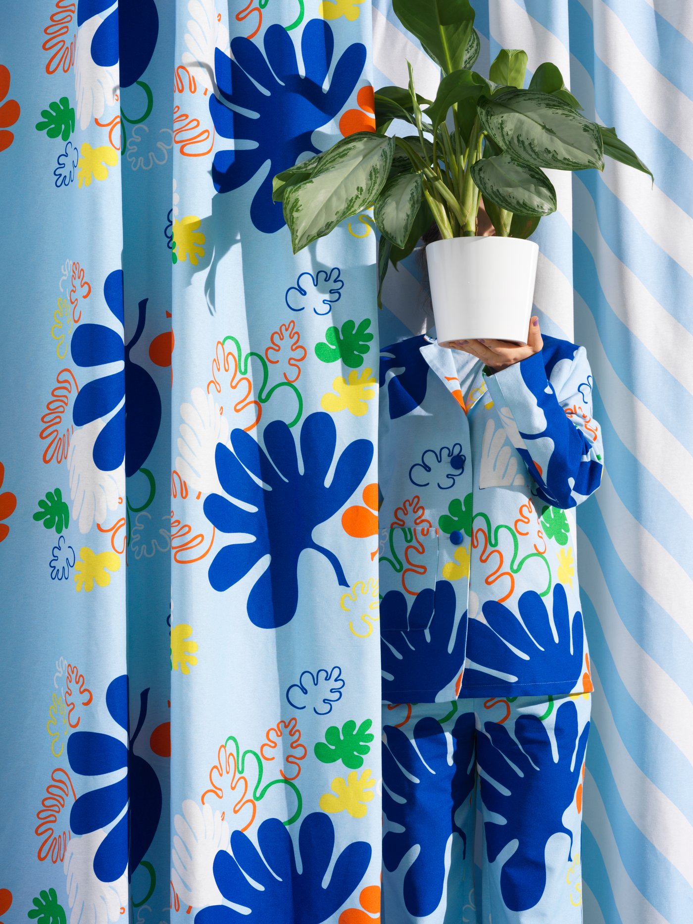 A person in a suit made from KRYPKORNELL pre-cut fabric emerges from matching curtains with a plant pot covering their face.