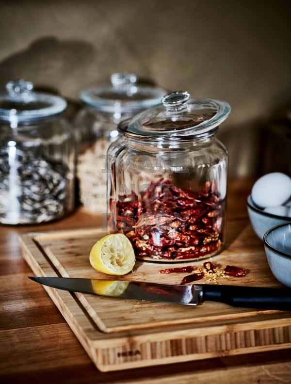 An IKEA VARDAGEN glass jar with lid in a traditional style, filled with red dry spices. A wooden chopping board sitting on a walnut worktop, with a lemon cut in half and a chef knife resting on the chopping board.